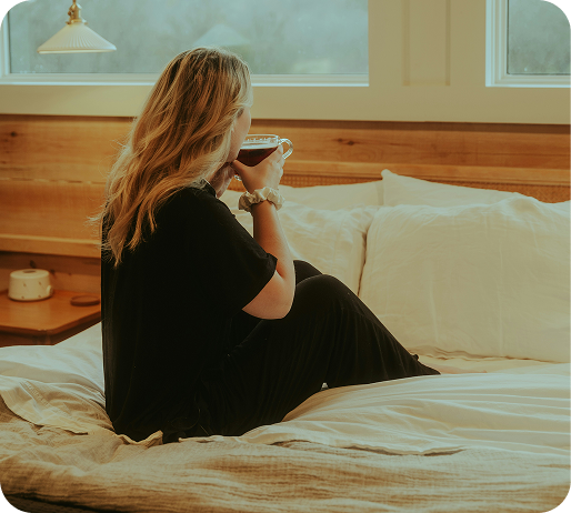 Woman sitting on bed drinking from a mug, reflecting quietly